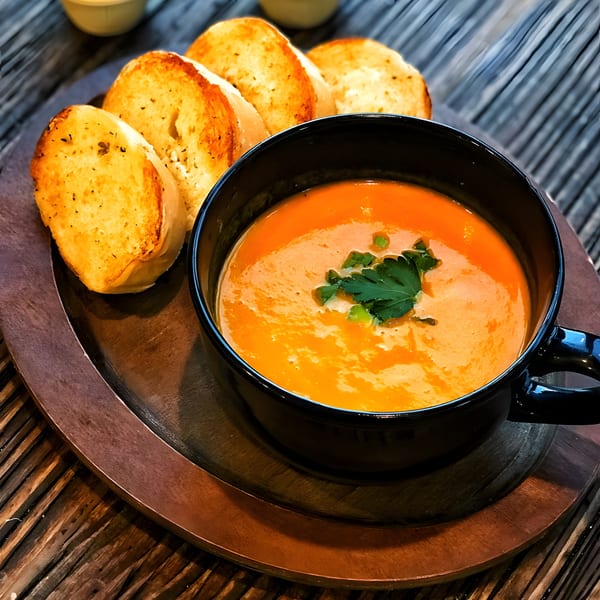 A black mug of vibrant orange Brazilian soup with parsley, served with toasted bread slices on a wooden tray.
