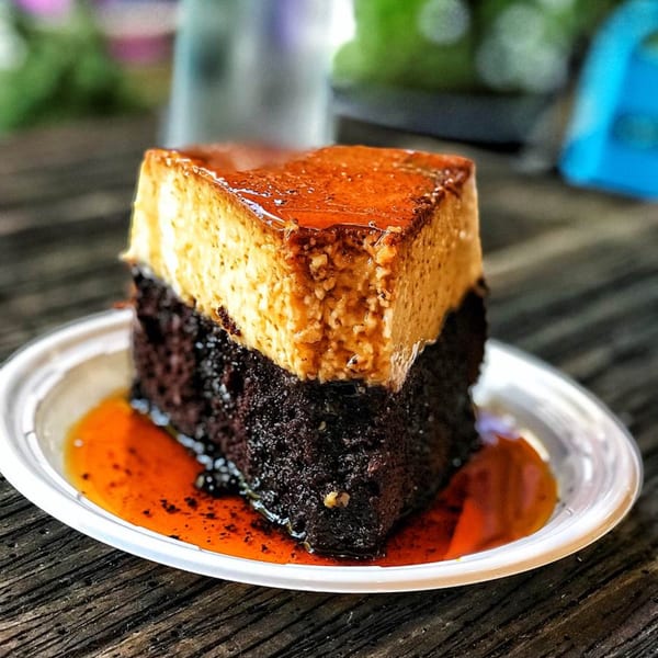 A slice of Brazilian chocolate cake with flan on top, drizzled with caramel sauce on a white plate.