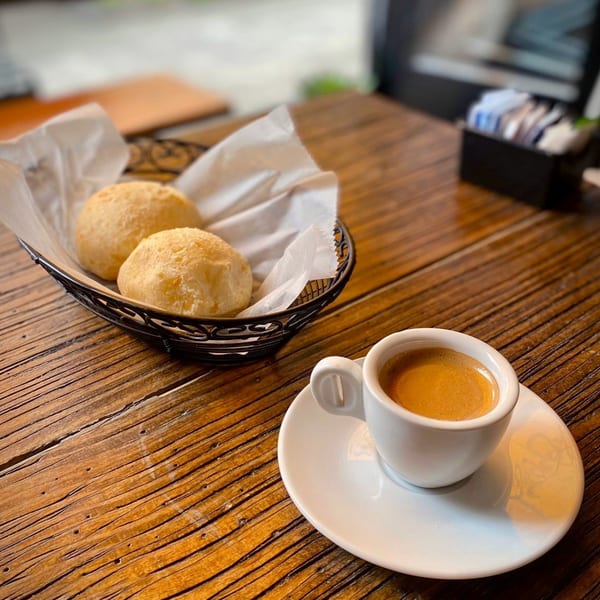 Two golden-brown Brazilian cheese breads in a basket with a cup of espresso on a wooden table.