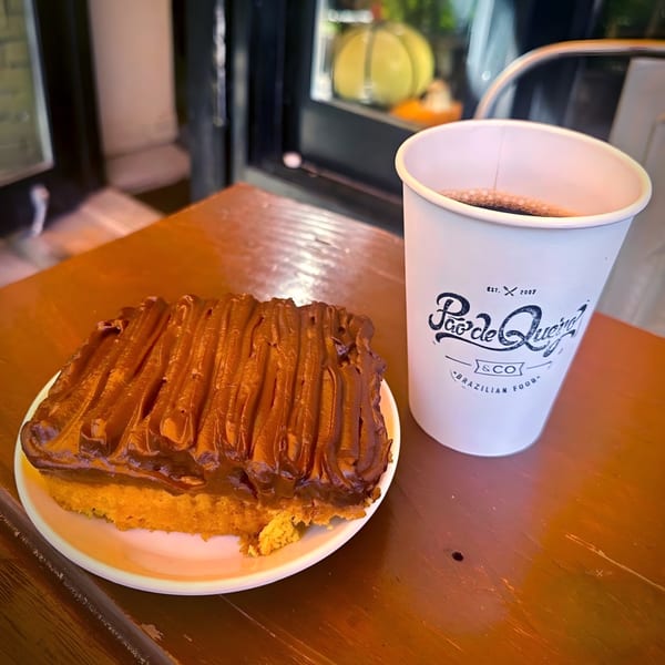 A slice of Brazilian carrot cake with chocolate frosting and a coffee cup on a wooden table.