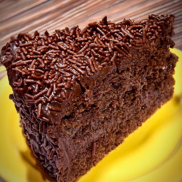 A slice of rich chocolate cake with chocolate frosting and sprinkles on a yellow plate.